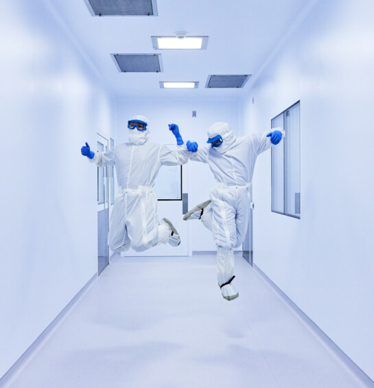 Two workers in white clean suits, goggles, and blue gloves jumping in the air.