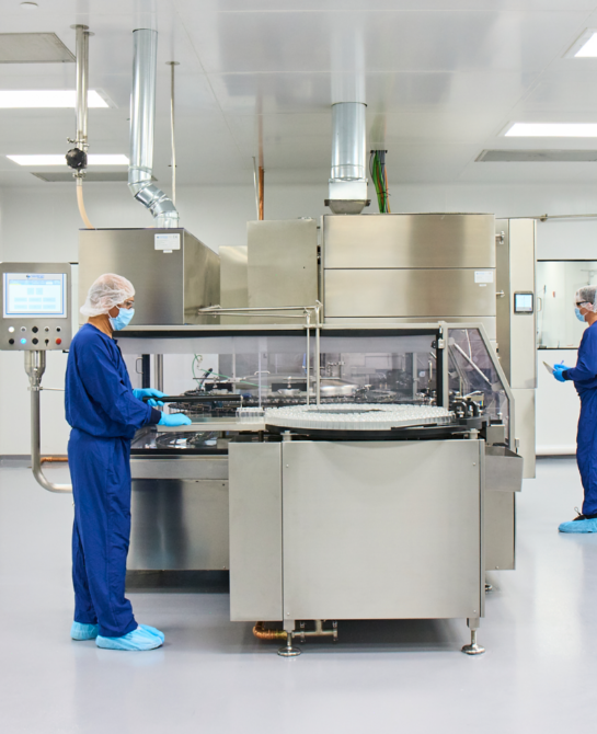 Two workers in blue scrubs, masks, and hairnets near a machine.