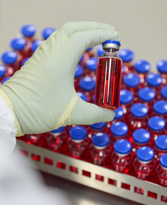 A hand holding a vial of red liquid with a tray full of vials in the background.