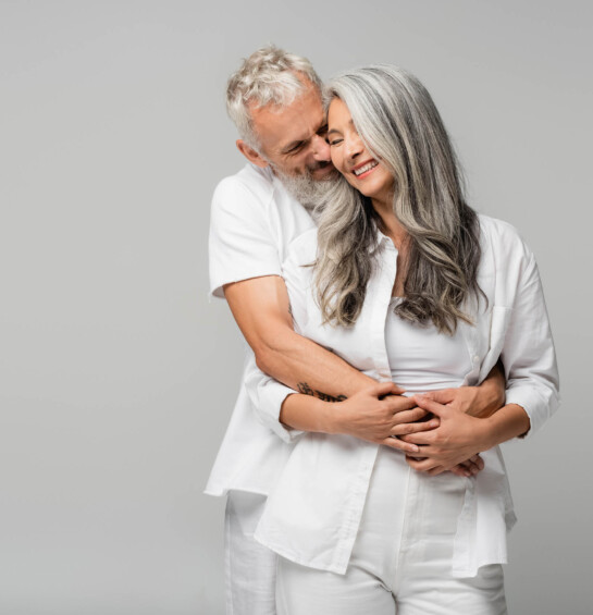 A older man hugging a smiling older woman from behind both wearing white